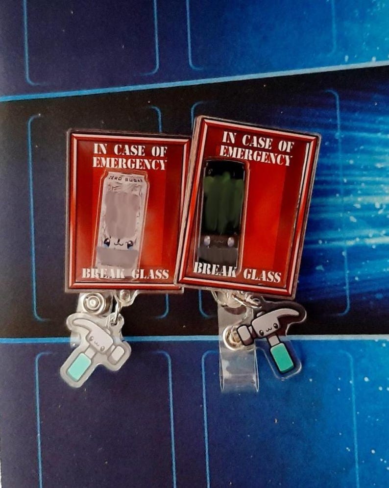 May include: Two novelty badge reels with the text "IN CASE OF EMERGENCY" and "BREAK GLASS". One reel features a clear glass design, the other a green glass design. Each has a hammer charm with a teal handle.