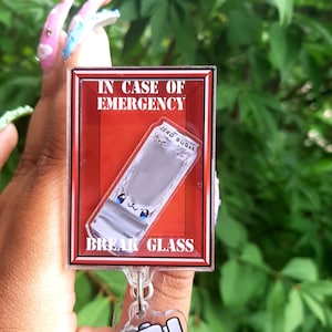 May include: A novelty acrylic keychain featuring a red box design with the text "IN CASE OF EMERGENCY" and "BREAK GLASS". Inside the box is a cartoon-style packet of "ZERO SUGAR". A small hammer charm is attached.