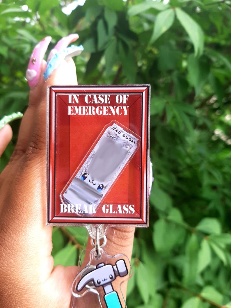 May include: A novelty acrylic keychain featuring a red box design with the text "IN CASE OF EMERGENCY" and "BREAK GLASS". Inside the box is a cartoon-style packet of "ZERO SUGAR". A small hammer charm is attached.