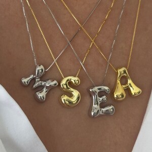 Könnte beinhalten: Vier goldene und silberne Halsketten mit Buchstabenanhängern, die das Wort "SEA" buchstabieren. Die Buchstaben sind in einer dicken, blasenartigen Schrift.