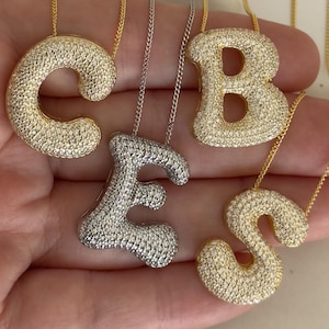 May include: Four gold and silver necklaces with letter pendants. The pendants are made of cubic zirconia and are shaped like the letters C, B, E, and S. The necklaces are on a white background.
