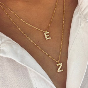 May include: Two gold necklaces with initial pendants. The top necklace has a pendant with the letter "E" and the bottom necklace has a pendant with the letter "Z". Both pendants are encrusted with small, clear stones.