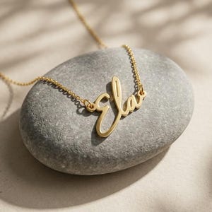 May include: A gold-colored necklace with the name "Ela" in a cursive font. The necklace is displayed on a gray stone, with the chain extending out of frame. The background is a neutral color with shadows.