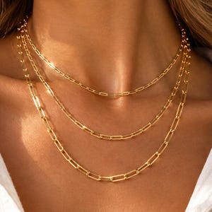 May include: A layered gold necklace set featuring three distinct chains with elongated rectangular paperclip links. The polished gold metal reflects light, creating a bright, metallic sheen across the interconnected chain links worn around the neck.