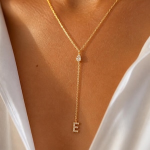 May include: A gold lariat necklace featuring a delicate chain, a single pear-shaped clear stone at the center, and a dangling letter E pendant encrusted with small clear stones at the bottom of the vertical drop chain.