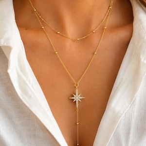 May include: A gold-toned layered necklace featuring a delicate chain with small evenly spaced beads. The lower layer includes a central starburst pendant encrusted with clear stones, with a thin chain extending downward from the starburst.