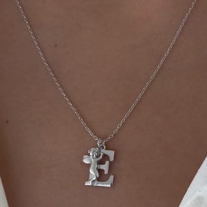 May include: A silver necklace with a pendant featuring a small angel holding the letter E.