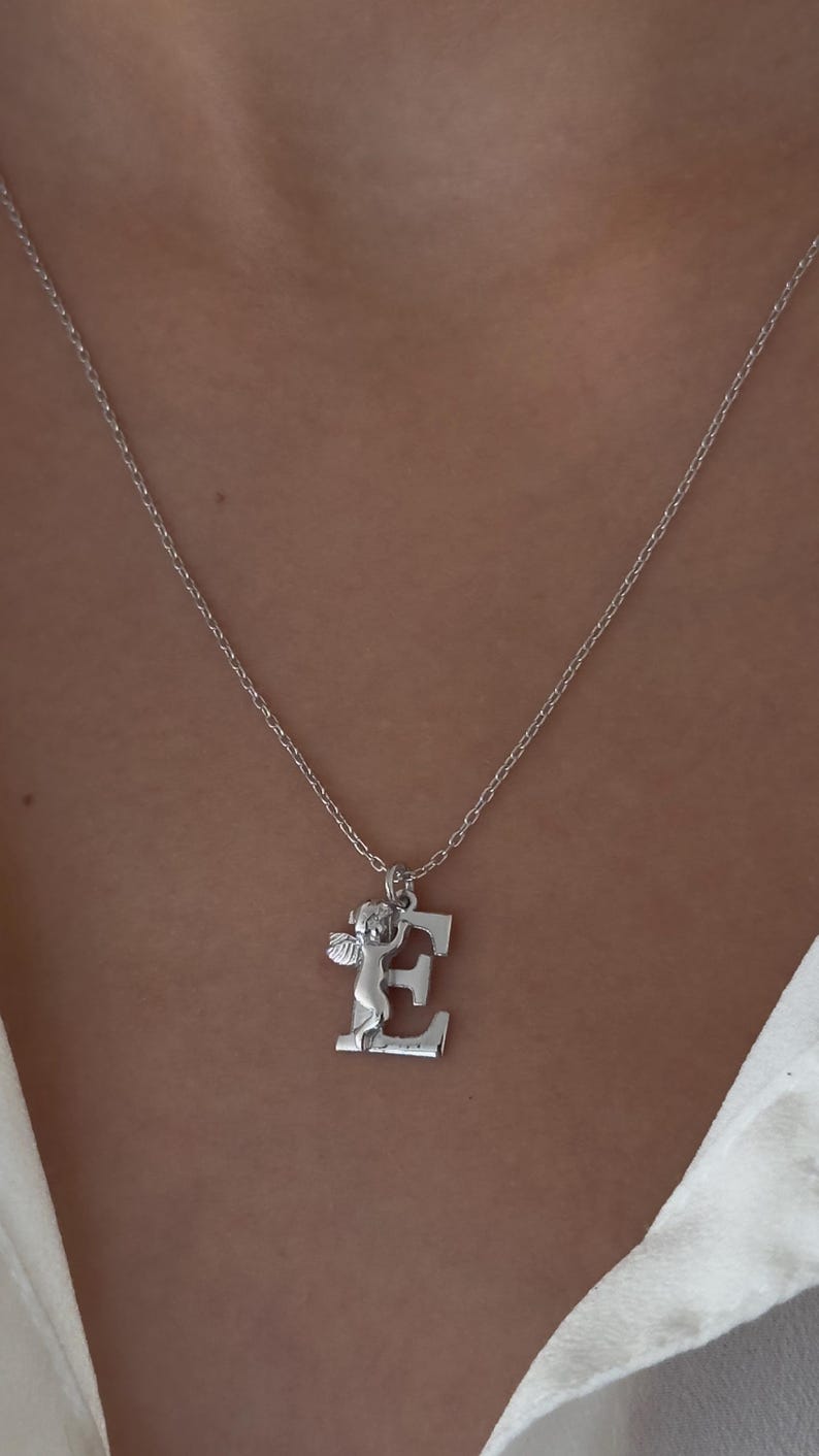 May include: A silver necklace with a pendant featuring a small angel holding the letter E.