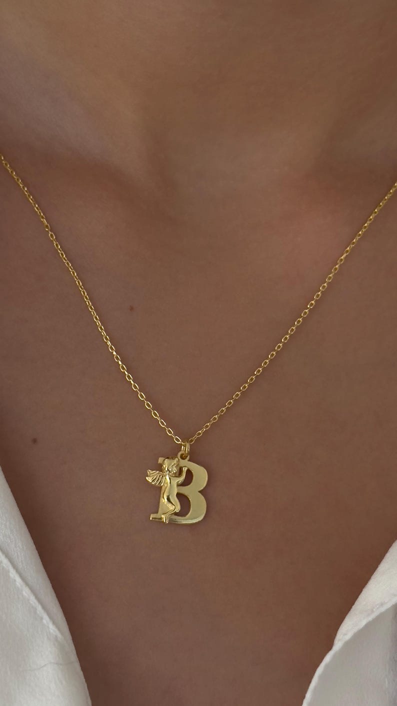 May include: A gold chain necklace with a pendant featuring the letter "B" with an angel figure on top.