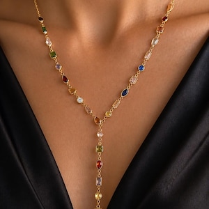May include: A gold Y-shaped necklace featuring a delicate chain adorned with a variety of multi-coloured gemstones in diverse shapes, including round, rectangular, and pear-cut stones in shades of blue, red, green, yellow, and clear white.