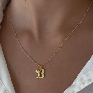 May include: A gold chain necklace with a pendant featuring the letter "B" and a small angel figure.