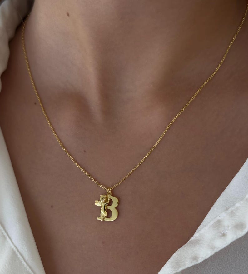 May include: A gold chain necklace with a pendant featuring the letter "B" and a small angel figure.
