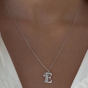 May include: A silver necklace with a pendant featuring the letter "E" with a small angel figure on it.