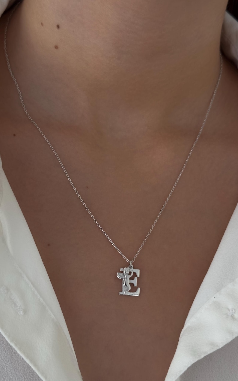 May include: A silver necklace with a pendant featuring the letter "E" with a small angel figure on it.