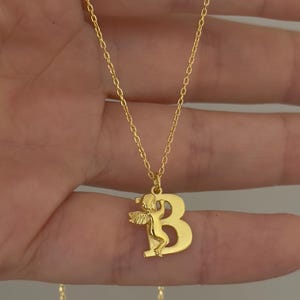 May include: Gold chain necklace with a pendant featuring the letter "B" with an angel figure inside.