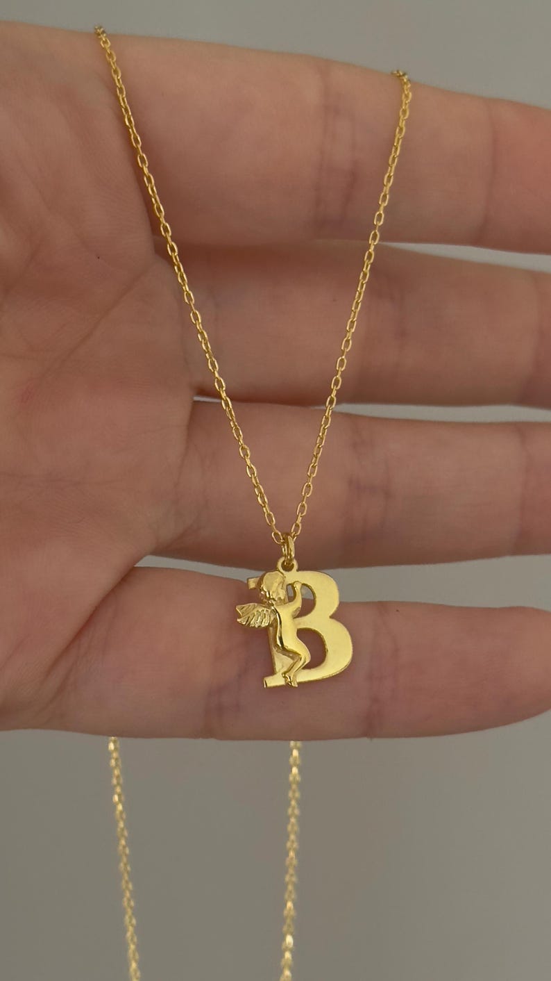 May include: Gold chain necklace with a pendant featuring the letter "B" with an angel figure inside.