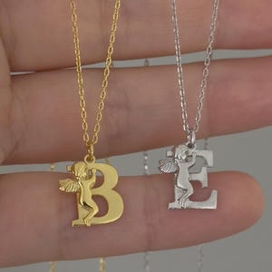May include: Two gold necklaces with angel wings and letter pendants. The necklace on the left has a gold chain and a letter "B" pendant. The necklace on the right has a silver chain and a letter "E" pendant.