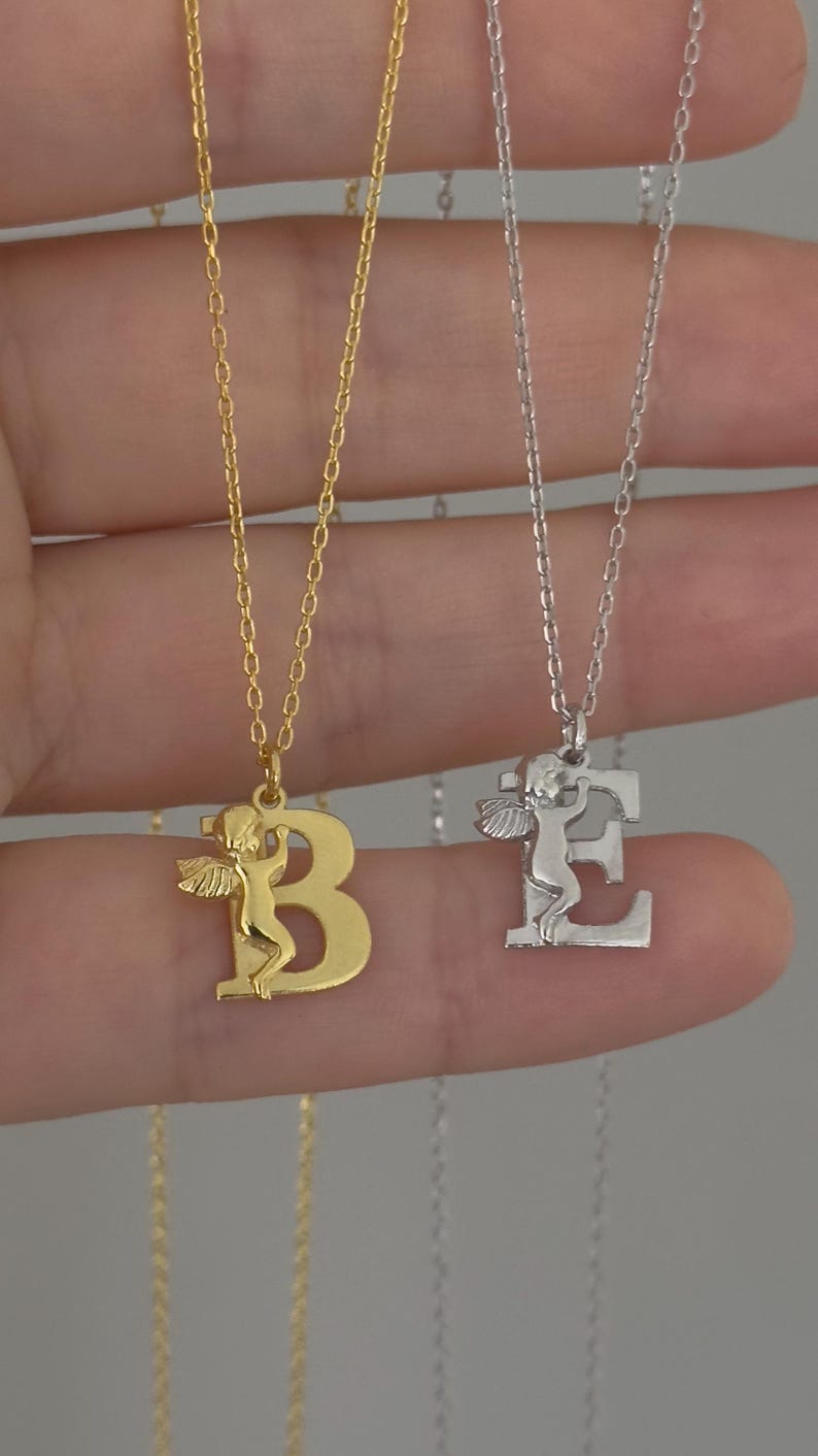 May include: Two gold necklaces with angel wings and letter pendants. The necklace on the left has a gold chain and a letter "B" pendant. The necklace on the right has a silver chain and a letter "E" pendant.
