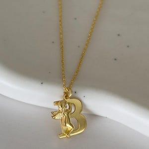 May include: A gold chain necklace with a pendant featuring the letter "B" with a small angel figure holding onto the letter.
