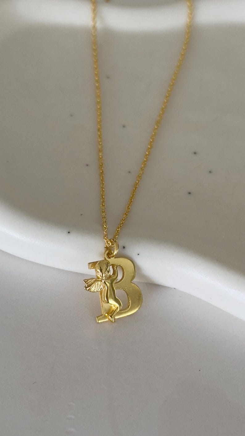 May include: A gold chain necklace with a pendant featuring the letter "B" with a small angel figure holding onto the letter.