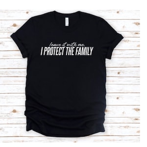 May include: Black t-shirt with the white text "leave it with me. I PROTECT THE FAMILY." The shirt has short sleeves and a crew neck. The background is a light wood grain.