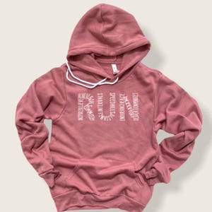 May include: A dusty rose hoodie with a white graphic that reads "RUN" with words vertically stacked around the letters. The words are: "Inspire", "Believe", "Commit", "Goals", "Max Intervals", "Sustain", "Speedhill", "Endurance", "Accept", "Embrace", "Strength", "Mindset".