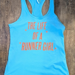 May include: A light blue racerback tank top with the text "THE LIFE OF A RUNNER GIRL" in orange glitter lettering. The shirt has a scoop neck and a racerback design, perfect for running.