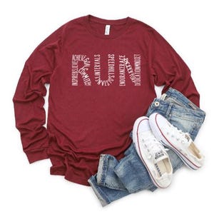 May include: A maroon long-sleeve shirt with the word "RUN" spelled out in white text. The letters are made up of words related to running, such as "achieve", "inspire", "believe", "commit", "strength", "intervals", "goals", "thinking", "speed", "endurance", "race", "effort", "believe", "dedication", and "mindset".