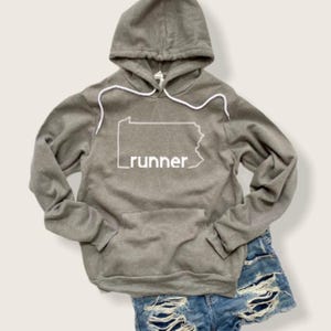 Puede incluir: Una sudadera con capucha gris con un contorno blanco del estado de Pensilvania y la palabra "runner" impresa en la parte delantera.