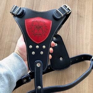 Könnte beinhalten: Schwarzes Lederhundgeschirr mit einem roten Patch, auf dem eine schwarze und weiße Hundesilhouette abgebildet ist. Das Geschirr hat silberne Schnallen und Metallnieten.