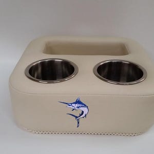 May include: Beige boat cup holder with two stainless steel cup holders and a rectangular storage area. A blue and white marlin fish graphic is on the front. The base has a decorative trim.