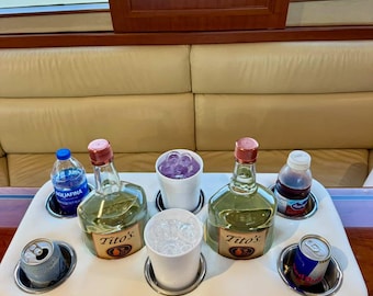 May include: A white, cushioned drink holder with stainless steel cup holders. It holds two bottles of Tito's vodka, two cups with ice, two cans, a bottle of water, and a small bottle of juice. The background shows a boat interior.