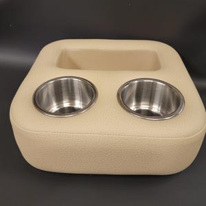 May include: Beige leatherette car console with two stainless steel cup holders and a rectangular storage area. The console is designed to fit in a vehicle and has a stitched edge.