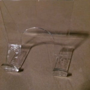 May include: A clear acrylic display stand with a curved top and a flat base. The stand is designed to hold a small object, such as a figurine or a collectible.