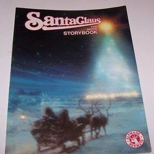 May include: A book cover with a blue and white illustration of Santa Claus flying in a sleigh pulled by reindeer. The book title is "Santa Claus The Movie Storybook" with a red and white Elf Made logo in the bottom right corner.