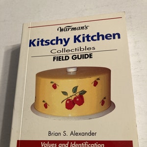 May include: A book titled "Kitsch Kitchen Collectibles Field Guide" by Brian S. Alexander. The book cover features a yellow cake stand with red apples and green leaves.