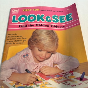 Könnte beinhalten: Ein Kind malt mit Buntstiften in einem "LOOK & SEE" Aktivitätsbuch. Das Buchcover ist rosa und trägt den Text "FIRST FUN preschool activities" und "Find the Hidden Objects". Das Kind trägt ein rosa Hemd.