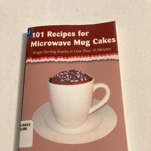 Könnte beinhalten: Ein Kochbuch mit dem Titel "101 Rezepte für Mikrowellen-Becherkuchen" zeigt eine weiße Tasse gefüllt mit einem Schokoladenkuchen, der mit roten, weißen und blauen Streuseln belegt ist. Das Buchcover ist braun und rosa.