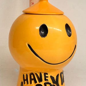 May include: A bright yellow ceramic jar with a lid, featuring a black smiley face and the text "HAVE A HAPPY" in black lettering. A wooden ruler is positioned behind the jar, showing the lid reaches approximately 11 inches in height.