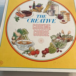 May include: A vintage cookbook titled "The Creative Cooking Course" with a yellow cover and a circular illustration. The illustration features various food items like meat, vegetables, and salads, with the title in blue and red.
