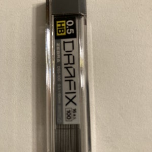 Puede incluir: Un contenedor de plástico transparente de 100 minas de lápiz mecánico Zebra DraFix. Las minas tienen un diámetro de 0,5 mm y están etiquetadas como HB. El contenedor es gris y tiene una etiqueta negra con texto blanco.