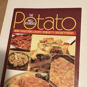 Peut inclure: Un livre de cuisine marron intitulé The Well-Rounded Potato avec le sous-titre Menu Suggestions & Recipes From Betty Crocker Potatoes. La couverture présente un collage de divers plats à base de pommes de terre, dont des gratins et salades.
