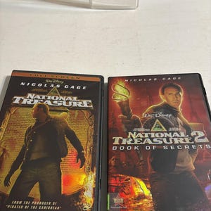 May include: Two DVD cases for the movies "National Treasure" and "National Treasure 2: Book of Secrets" starring Nicolas Cage. The first case has a brown and gold design, while the second is red and gold. Both cases feature the Walt Disney logo.