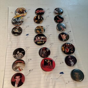 Könnte beinhalten: Eine Sammlung von einundzwanzig runden Ansteckbuttons auf linierten Papier, die verschiedene Musikkünstler, Bands und Filmthemen zeigen. Die Buttons weisen gedruckte Grafiken, Porträts und Texte auf, darunter Dio, Rocky IV, Y&T und Huey Lewis.
