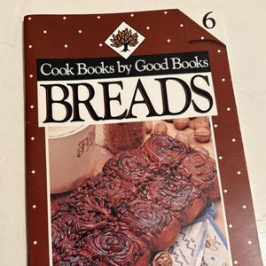 May include: A softcover cookbook titled Cook Books by Good Books BREADS, featuring the number 6 in the top right corner. The cover displays a photograph of dark, glazed cinnamon rolls topped with nuts, alongside a mug and scattered walnuts.