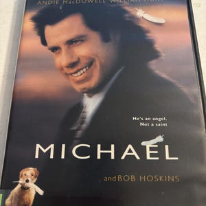 Puede incluir: Caja de DVD de la película "Michael" con John Travolta, Andie MacDowell y William Hurt. La portada muestra a Travolta sonriendo, un gráfico de pluma y el texto "He's an angel. Not a saint."