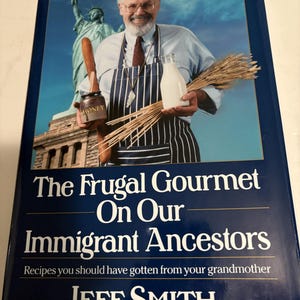 May include: A hardcover book titled The Frugal Gourmet On Our Immigrant Ancestors by Jeff Smith. The cover features a man in a blue shirt and striped apron holding a honey jar, milk bottle, and wheat stalks, with the Statue of Liberty in the background.