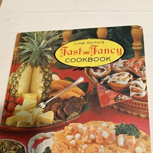 以下が含まれることがあります： June Roth's Fast and Fancy Cookbookというタイトルのハードカバーの料理本。表紙には、パイナップル丸ごと、パイナップルのスライス、ダークミート、銀のフォーク、シナモンロール、白のトッピングがのったエビ料理など、色鮮やかな料理のコラージュが描かれています。