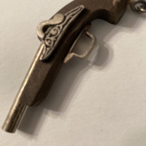 Op de afbeelding: Een klein, zilverkleurig speelgoedpistool met een bruin houten handvat. Het pistool heeft een decoratief ontwerp aan de zijkant.