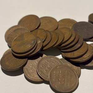 May include: A collection of bronze-colored United States one-cent coins, also known as pennies. The coins are scattered, with some stacked, and show the text "ONE CENT". The coins have a worn appearance.